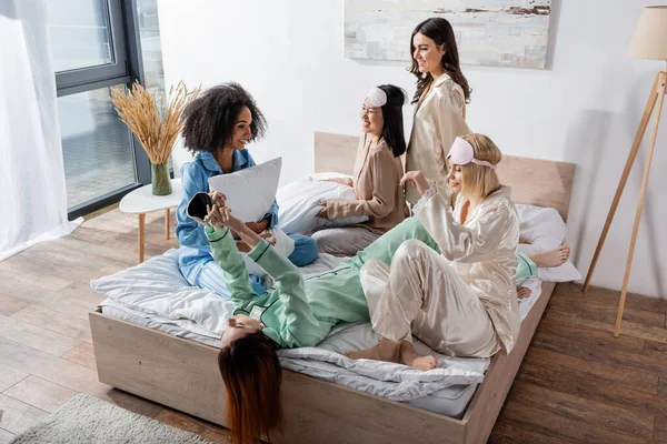 Groupe d'amis interracial souriants en pyjama assis sur le lit pendant la soirée pyjama — Photo de stock