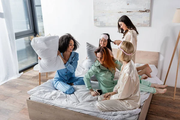 Juguetones amigos interracial en pijama teniendo pelea de almohadas durante la fiesta de pijamas - foto de stock