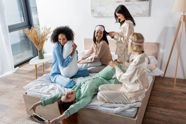 Улыбающиеся межрасовые друзья в пижаме, сидящие на кровати во время пижамной вечеринки — стоковое фото