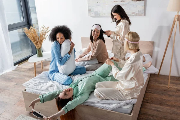 Sonrientes amigos interracial en pijama sentado en la cama durante la fiesta de pijamas - foto de stock