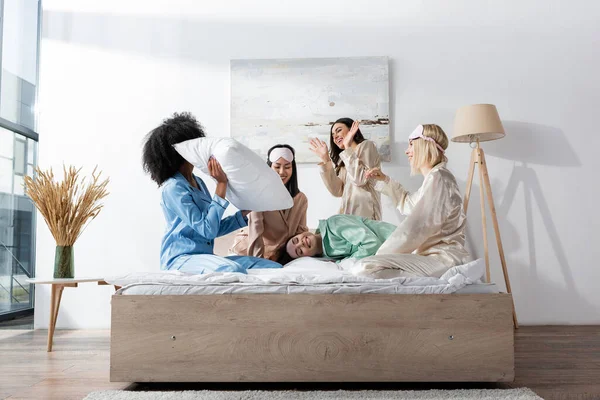 Smiling interracial friends in pajamas having pillow fight during slumber party — Stock Photo