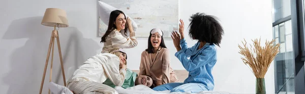 Група веселих міжрасових друзів у піжамі, що борються подушкою під час вечірки з димом, банер — стокове фото