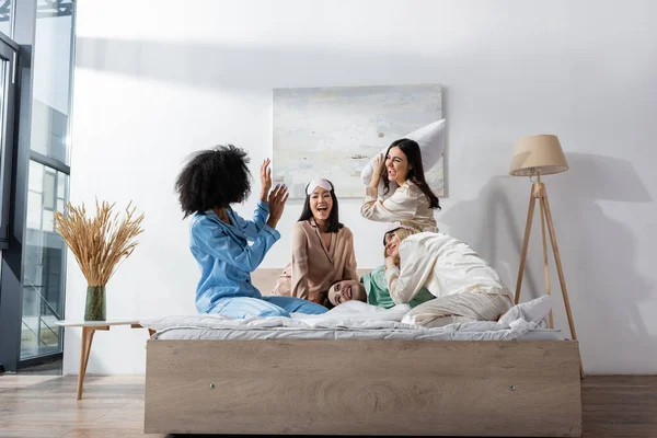 Group of amazed and happy interracial friends in pajamas having pillow fight during slumber party — Stock Photo