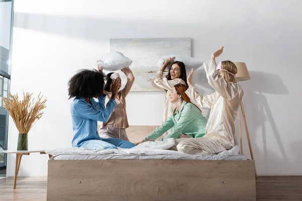 Веселые юные межрасовые друзья в пижаме дерутся на подушках во время пижамной вечеринки — стоковое фото