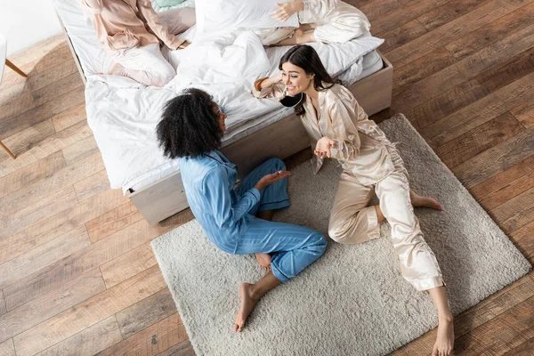 Vue grand angle de la femme afro-américaine assise sur le tapis et parlant avec un ami gai pendant la soirée pyjama — Photo de stock