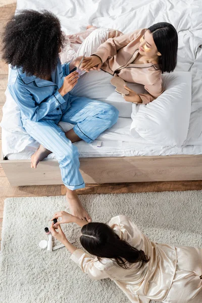 Vista superior de la mujer afroamericana haciendo manicura a amigo asiático durante la fiesta de pijamas - foto de stock