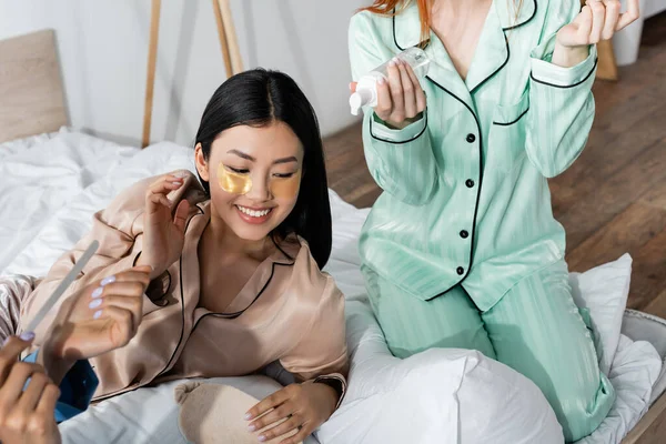 Cheerful asian woman with eye patches smiling near friends during slumber party — Stock Photo