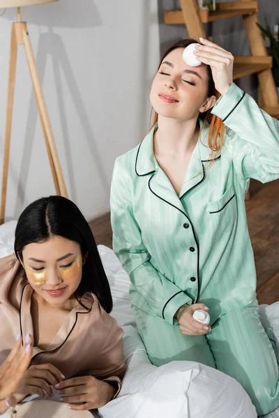 Feliz pelirroja mujer la eliminación de maquillaje con algodón almohadilla cerca asiático amigo - foto de stock