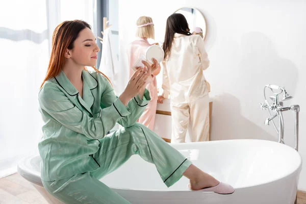 Руда жінка тримає контейнер з косметичним кремом біля розмитих друзів у ванній — стокове фото