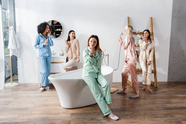 Cheerful interracial women holding cosmetic products in bathroom during slumber party — Stock Photo