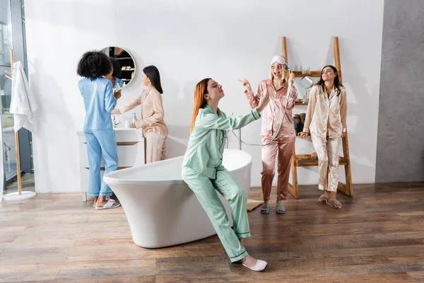 Redhead woman spraying mist on face near interracial friends in bathroom — Stock Photo