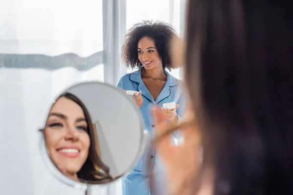 Feliz Africano americano mulher segurando recipiente com creme cosmético perto amigo borrado sorrindo em primeiro plano — Fotografia de Stock