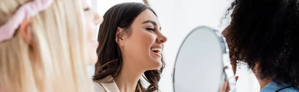 Happy interracial friends near blonde young woman with mirror on blurred foreground, banner — Stock Photo