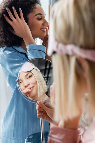 Mujer rubia feliz en la máscara del sueño mirando el espejo cerca de un amigo afroamericano - foto de stock