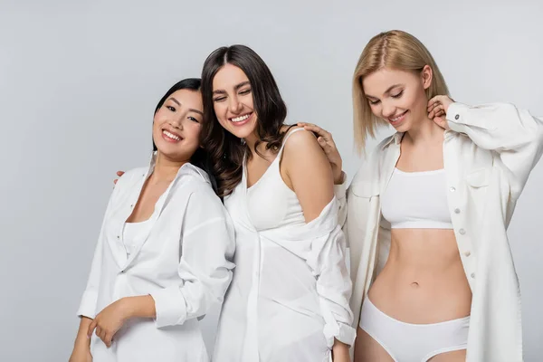 Joyful interracial young models in white shirts smiling isolated on grey — Stock Photo