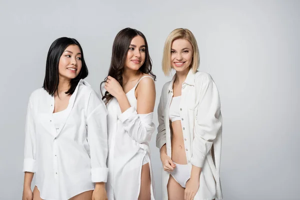 Fröhliche multiethnische junge Models in weißen Hemden lächeln vereinzelt auf grau — Stockfoto