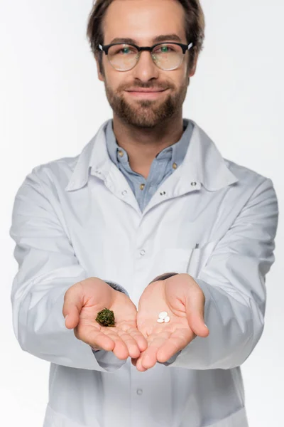 Médico sonriente con cannabis medicinal seco y pastillas en las manos aisladas en blanco - foto de stock