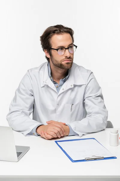 Doctor sentado cerca de portapapeles, portátil y contenedor con pastillas aisladas en blanco, concepto de cannabis medicinal - foto de stock