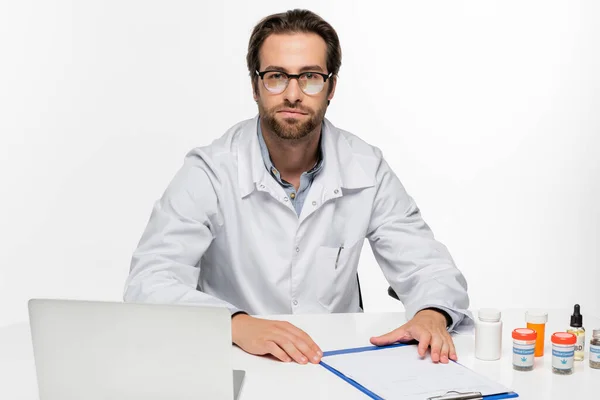 Arzt blickt auf Kamera in der Nähe von Behältern mit medizinischem Hanf, Laptop und Klemmbrett isoliert auf weiß — Stockfoto