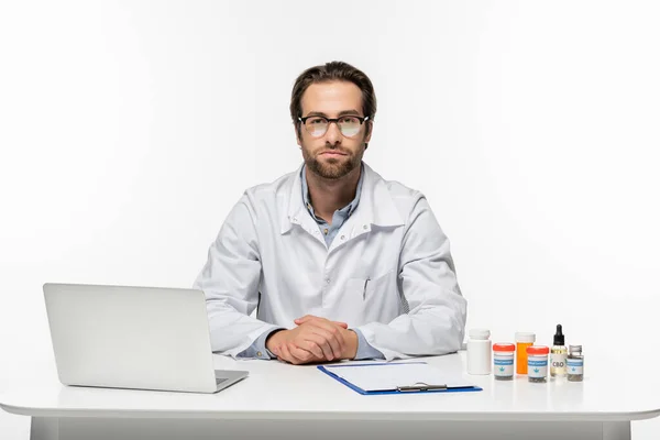 Doctor in eyeglasses near laptop, clipboard and medical cannabis medication isolated on white — Stock Photo