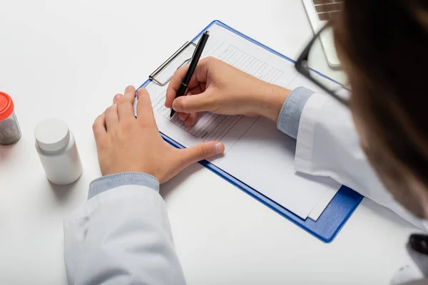 Partial view of blurred physician writing on clipboard near containers with treatment, medical cannabis concept — Stock Photo