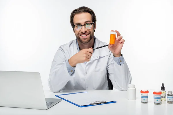 Médecin souriant pointant avec stylo au récipient avec des pilules de cannabis médical isolé sur blanc — Photo de stock