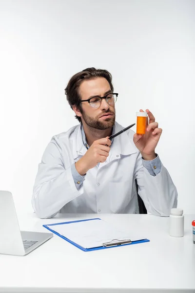 Doctor pointing with pen at container with medical cannabis pills near clipboard and laptop isolated on white — Stock Photo