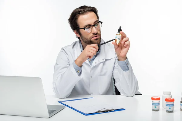 Medico indicando fiala di olio di canapa vicino appunti e laptop isolato su bianco — Foto stock