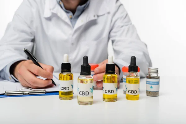 Cropped view of doctor writing prescription near bottles with cbd oil isolated on white — Stock Photo