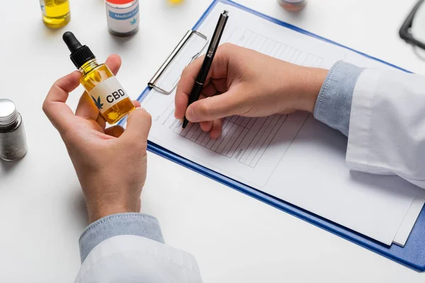 Partial view of doctor with vial of cbd oil writing prescription on clipboard — Stock Photo