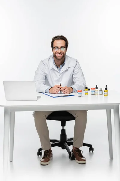 Щасливий лікар сидить за столом з ноутбуком і контейнерами з медичним канабісом на білому — стокове фото