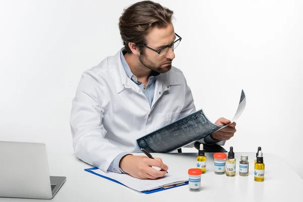 Medico che scrive prescrizione mentre tiene la risonanza magnetica vicino a contenitori con cannabis medica isolata su bianco — Foto stock
