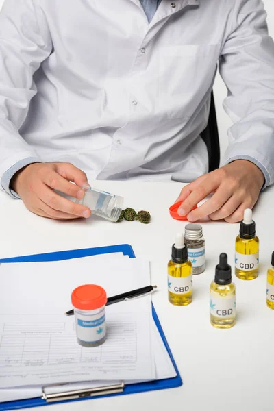 Vista ritagliata del medico vicino a cannabis medica secca e contenitori con trattamento isolato su bianco — Foto stock