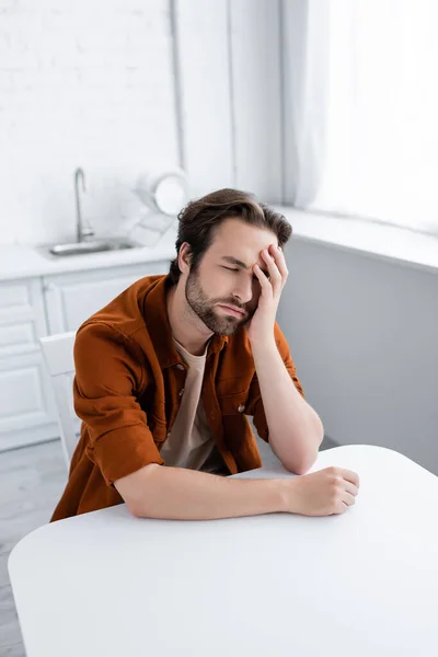 Hombre sentado en la mesa de la cocina y sufriendo de migraña - foto de stock