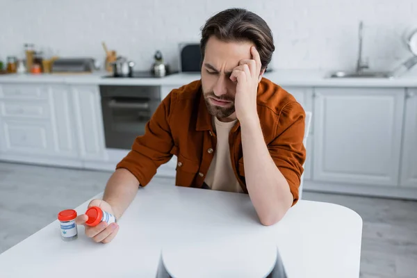 Hombre que sufre de dolor de cabeza mientras mira contenedor con cannabis medicinal en la cocina - foto de stock