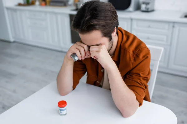Homem doente cobrindo os olhos com punhos enquanto segurando recipiente com cannabis medicinal na cozinha — Fotografia de Stock