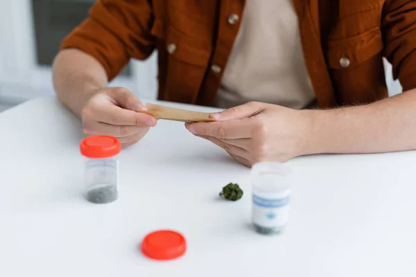 Teilansicht eines kranken Mannes, der Joint mit medizinischem Hanf in der Nähe von Behältern auf dem Tisch macht — Stockfoto