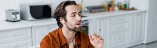 Man smoking joint of medical cannabis in kitchen, banner — Stock Photo