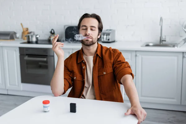 Homem fumando cannabis medicinal perto frasco borrado e isqueiro em casa — Fotografia de Stock