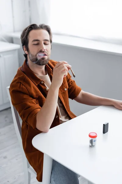 Smoker holding cigarette of medical cannabis near lighter and jar on table — Stock Photo