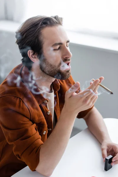 Homem segurando mais leve e articulação de cannabis medicinal perto fumaça turva — Fotografia de Stock