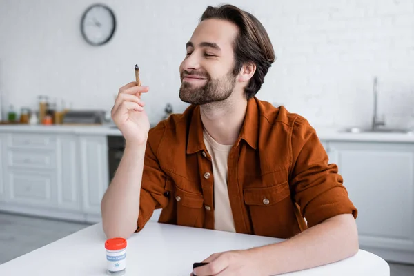 Hombre sonriente mirando la articulación de cannabis medicinal cerca del frasco en la mesa - foto de stock