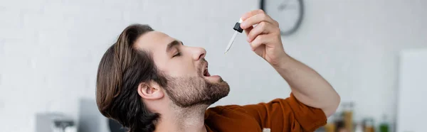 Vista lateral del hombre sosteniendo la pipeta con aceite de cbd cerca de la boca abierta, pancarta - foto de stock