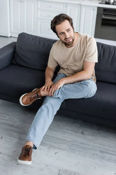 Müder Mann berührt Bein, während er Schmerzen auf der Couch spürt — Stockfoto