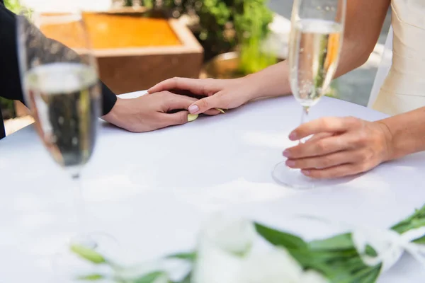 Обрезанный вид лесбийской пары с размытым шампанским, держащимся за руки во время свадьбы на открытом воздухе — стоковое фото