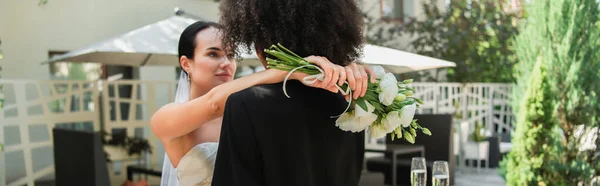 Jovem lésbica no vestido segurando buquê enquanto abraça a namorada afro-americana durante o casamento, banner — Fotografia de Stock