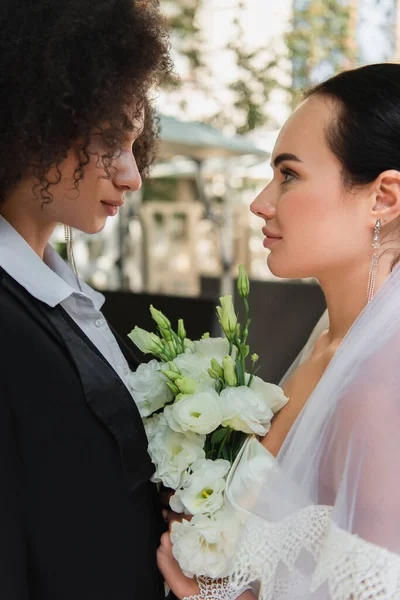 Vista lateral de mujeres lesbianas interracial con ramo mirándose durante la boda al aire libre - foto de stock