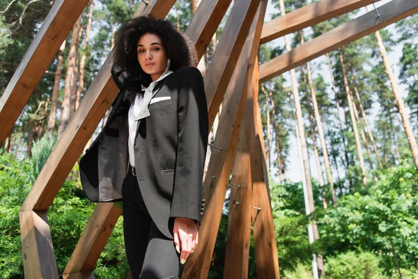 Low angle view of african american woman in formal wear looking at camera near wooden arch — Stock Photo