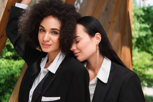 Woman in formal wear standing with closed eye near african american girlfriend in park — Stock Photo