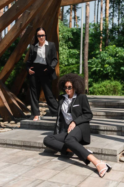 Femme afro-américaine en tenue formelle assise près de la petite amie et arche dans le parc — Photo de stock